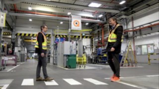 Zwei Mitarbeiter begegnen sich im Lager, während sie die Linde Secure Distance Vest und den Linde Distance Beeper tragen. Zwei Mitarbeiter begegnen sich im Lager, während sie die Linde Secure Distance Vest und den Linde Distance Beeper tragen.