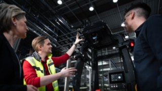 Ein Mitarbeiter von Linde MH steht vor einem automatisierten Fahrzeug und erklärt zwei Kunden etwas darüber