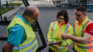 Drei Personen mit Warnwesten im Gespräch: Sicherheitsberatung vor Ort beim Kunden. Drei Personen mit Warnwesten im Gespräch: Sicherheitsberatung vor Ort beim Kunden.