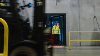 Ein Mann in Sicherheitskleidung tritt aus einer Tür in eine Werkshalle. Ein Stapler fährt vorbei. Ein Mann in Sicherheitskleidung tritt aus einer Tür in eine Werkshalle. Ein Stapler fährt vorbei.