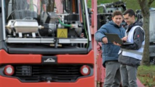 Autonome Fahrzeuge müssen genauso aufmerksam, reaktionsschnell, vorausschauend agieren wie menschliche Staplerfahrer. Zwei Männer stehen vor einem autonomen Stapler und schauen gemeinsam auf die Daten am Laptop.