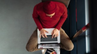 Mann mit roter Mütze und rotem Pulli sitzt im Schneidersitz am Laptop