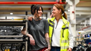 Zwei Mitarbeitende im Gespräch in einer Produktionsumgebung mit technischen Anlagen im Hintergrund