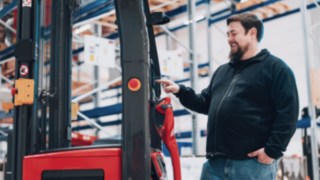 Ein lachender Mann in einer Lagerhalle konfiguriert ein Gerät von Linde, indem er mit dem Zeigefinger auf einen roten Knopf drückt. Er trägt eine schwarze Fleecejacke und Jeans. Im Hintergrund sind Regale mit blauen Kisten zu sehen.