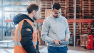 Zwei Mitarbeitende im technischen Austausch in einem Lager- oder Industrieumfeld, von denen einer eine Warnweste trägt und der andere ein Steuergerät bedient, was internationale Projekteinsätze, technische Verantwortung und globale Karrieremöglichkeiten symbolisiert.
