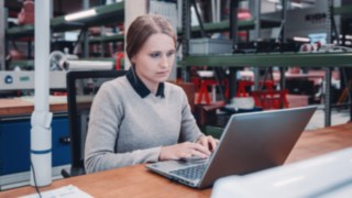 Eine Mitarbeiterin arbeitet konzentriert am Laptop in einer Werkstattumgebung, was symbolisch für berufliche Weiterbildung und persönliche Entwicklung bei KION Automation steht.