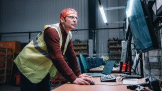 Ein Mitarbeiter mit Sicherheitsweste arbeitet konzentriert an einem Bildschirmarbeitsplatz in der Halle, was ein Symbol für das innovative und technologisch geprägte Arbeitsumfeld bei KION Automation ist.