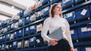 Eine Projektmanagerin mit Laptop in einer modernen Lagerhalle engagiert sich für nachhaltige und effiziente Lieferketten bei KION Automation.