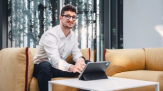 Ein junger Vertriebsmitarbeiter arbeitet mit einem Tablet in einer modernen Lounge-Umgebung, was für flexibles, kundenorientiertes Arbeiten im Vertrieb und den Einsatz digitaler Tools zur Präsentation innovativer Lösungen steht. 