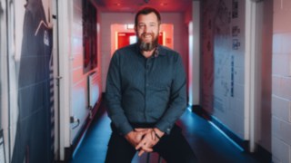 Ein Vertriebsmitarbeiter sitzt entspannt in einem markant ausgeleuchteten Unternehmensflur und blickt lächelnd in die Kamera, was für Persönlichkeit, Kundennähe und authentisches Engagement im internationalen Vertrieb steht. 