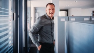 Ein lachender Vertriebsmitarbeiter in moderner Büroumgebung lebt die Vielfalt und Dynamik im Sales-Alltag mit ständig neuen Kundenkontakten, Herausforderungen und Lösungsansätzen. 