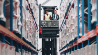 Zwei KION Automation-Mitarbeitende stehen in einem Hochregallager auf einem Schmalgangstapler zwischen Palettenreihen, was ein innovatives und dynamisches Arbeitsumfeld symbolisiert.