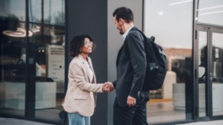 Zwei Geschäftsleute begrüßen sich mit einem Handschlag vor einem modernen Bürogebäude, was internationale Zusammenarbeit, kulturelle Offenheit und die globalen Karrieremöglichkeiten innerhalb eines vernetzten Unternehmensumfelds symbolisiert.