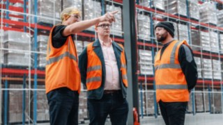 Drei Mitarbeitende in Warnwesten führen ein Gespräch in einem modernen Hochregallager, was teamorientierte Zusammenarbeit, fachlichen Austausch und eine kollaborative Unternehmenskultur in einem dynamischen Arbeitsumfeld verkörpert.