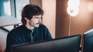 Ein Kollege aus dem Bereich Solution Design bei KION Automation arbeitet konzentriert an seinem Arbeitsplatz vor zwei Monitoren, umgeben von einer modernen Stehleuchte und Tageslicht. 