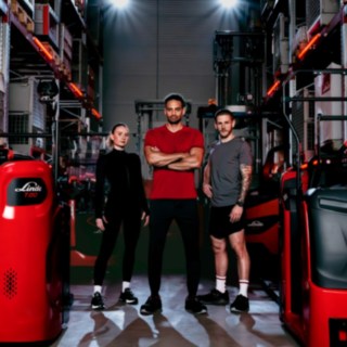 Eine Frau und zwei Männer in Sportkleidung stehen in einem Lager inmitten von Fahrzeugen von Linde Material Handling.